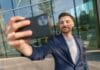 Efekt instagramowego przedsiębiorcy, czyli dlaczego młodzi chcą firmy dla zdjęć, a nie dla bilansu zysków i strat po roku Uśmiechnięty mężczyzna robi selfie smartfonem przed biurowcem