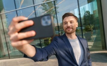 Efekt instagramowego przedsiębiorcy, czyli dlaczego młodzi chcą firmy dla zdjęć, a nie dla bilansu zysków i strat po roku Uśmiechnięty mężczyzna robi selfie smartfonem przed biurowcem