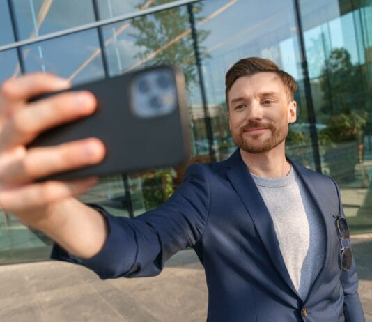 Efekt instagramowego przedsiębiorcy, czyli dlaczego młodzi chcą firmy dla zdjęć, a nie dla bilansu zysków i strat po roku Uśmiechnięty mężczyzna robi selfie smartfonem przed biurowcem